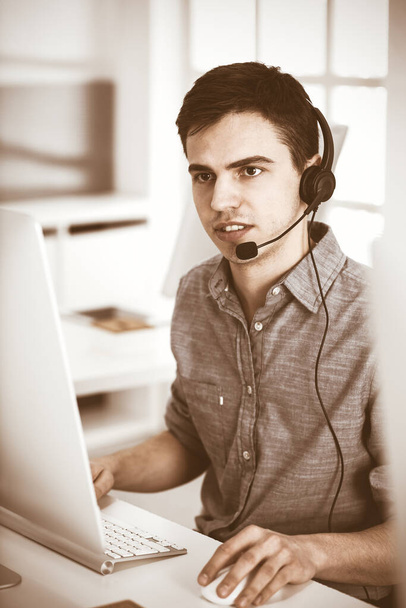 Casual dressed young man using headset and computer while talking with customers online. Call center, business concept - Foto, Imagem
