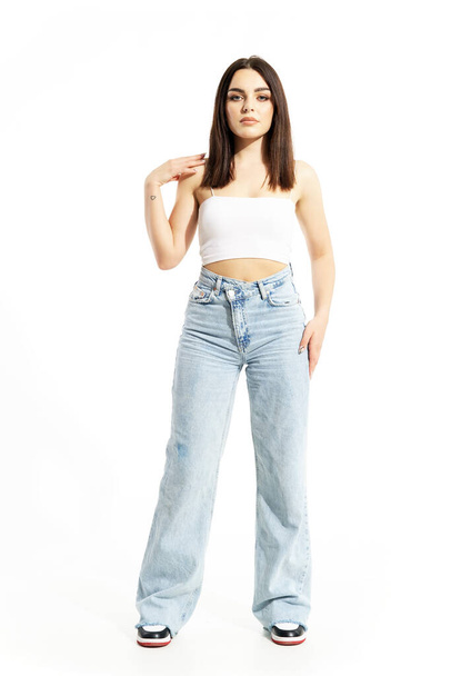 Gen Z feminine junge selbstbewusste Frau, die in weißem Top und Jeans mit berührender Schulter posiert. Volle Körperlänge isoliert auf weißem Hintergrund - Foto, Bild