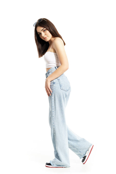 Kot pantolonlu ve kameraya bakan sevimli, modaya uygun Z jenerasyonlu kızın yan görüntüsü. Beyaz arkaplanda tam vücut uzunluğu izole edildi - Fotoğraf, Görsel