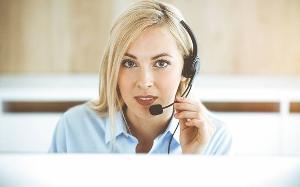 Blonde business woman sitting and communicated by headset in call center in sunny office. Concept of telesales business - Valokuva, kuva
