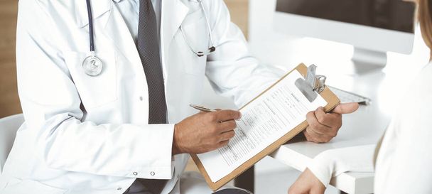 Doctor and patient discussing medical exam results while sitting at the desk in clinic, close-up. Male physician using clipboard for filling up medication history record of young woman - Foto, immagini