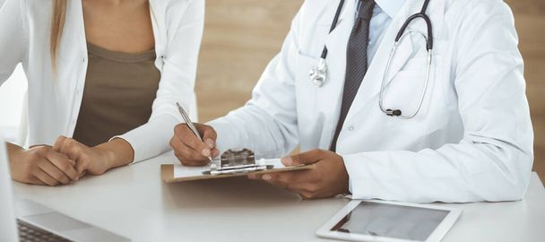 Doctor and patient discussing medical exam results while sitting at the desk in clinic, close-up. Male physician using clipboard for filling up medication history record of young woman - Foto, Imagen