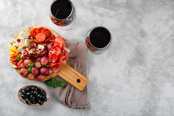  A set of appetizers for wine, jamon, pepperoni, cheese, grapes, peach and olives on a wooden board top view. Snack board and two glasses of red wine on light gray background, free space for text - Foto, imagen