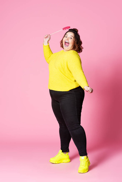 Happy plus size model in casual clothes with big lollipop, fat woman in black jeans, yellow jumper and sneakers on pink background, body positive concept - Foto, Bild