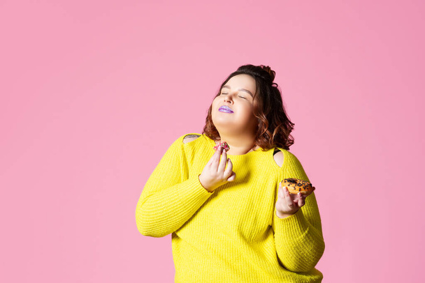 Happy plus size model eten donuts, dikke vrouw eet te veel en geniet op roze achtergrond, genieten van voedsel concept - Foto, afbeelding