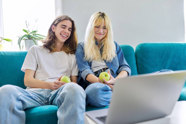 Laughing couple of teenagers having fun, sitting on couch, looking at laptop - Foto, imagen