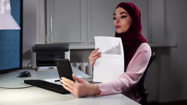 Femme musulmane dans le hijab fait un appel vidéo assis au bureau et montrant des documents avec des graphiques - Séquence, vidéo