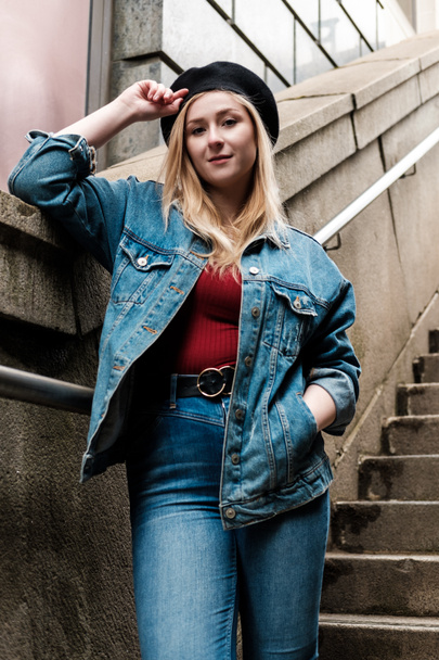 Jonge blonde vrouw poseert op de trap in baret, denim jas en broek en rood t-shirt. Ze staat op de trap.. - Foto, afbeelding