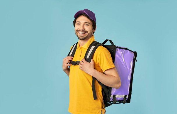Retrato de un joven mensajero, repartidor de uniforme con una mochila térmica aislada sobre un fondo azul. Entrega rápida a domicilio. Pedido en línea. - Foto, imagen