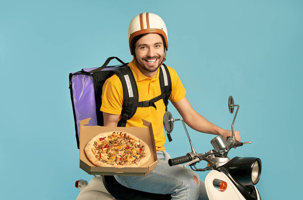 Junger Kurier, Pizzabote in Uniform mit Thermo-Rucksack auf einem Moped vor blauem Hintergrund. Schneller Transport per Expresslieferung nach Hause. Online-Bestellung. - Foto, Bild