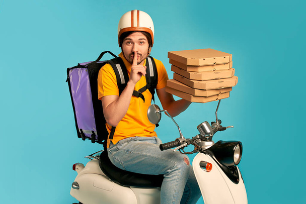 Young courier, pizza delivery man in uniform with thermo backpack on a moped isolated on blue background. Fast transport express home delivery. Online order. - Foto, imagen