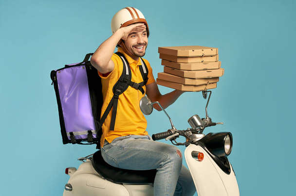 Young courier, pizza delivery man in uniform with thermo backpack on a moped isolated on blue background. Fast transport express home delivery. Online order. - Photo, Image