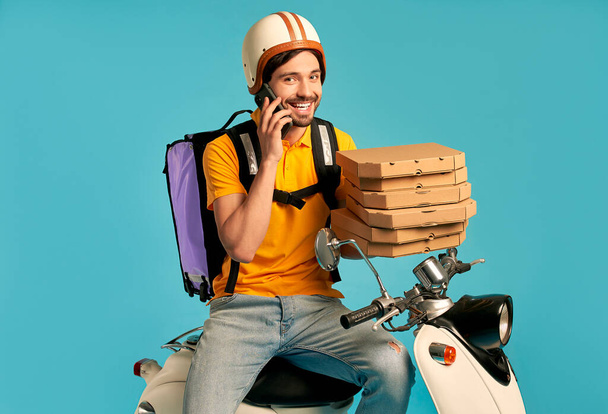 Mladý kurýr, poslíček pizzy v uniformě s termobatohem a telefonem na mopedu izolovaném na modrém pozadí. Rychlá doprava expresní doručení domů. Online objednávka. - Fotografie, Obrázek