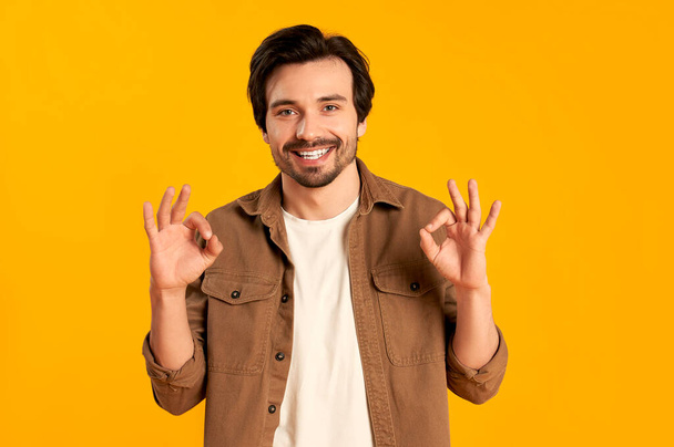 Young bearded man shows a gesture with his hands all ok against an orange background. - Photo, Image