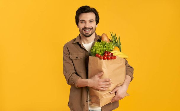 Young bearded man with paper package of vegetables groceries isolated on orange background. - Photo, image