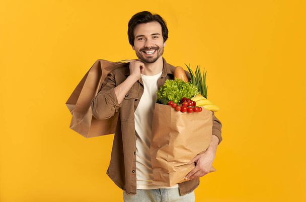 Junger bärtiger Mann mit Papiertüte mit Gemüseeinkäufen und Einkäufen isoliert auf orangefarbenem Hintergrund. - Foto, Bild