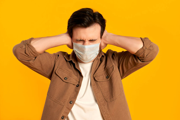 Young bearded man in protective medical mask covering his ears with his hands isolated on orange background. Life in the quarantine of COVID-19 coronavirus infection. - Fotografie, Obrázek
