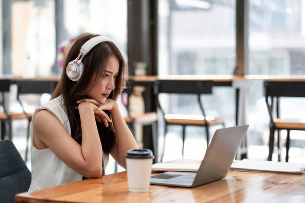Концентрована молода азіатка в навушниках дивиться на ноутбук для онлайн-навчання, приваблива мова навчання студенток, дивиться онлайн-вебінар, концепція освіти електронного навчання
. - Фото, зображення