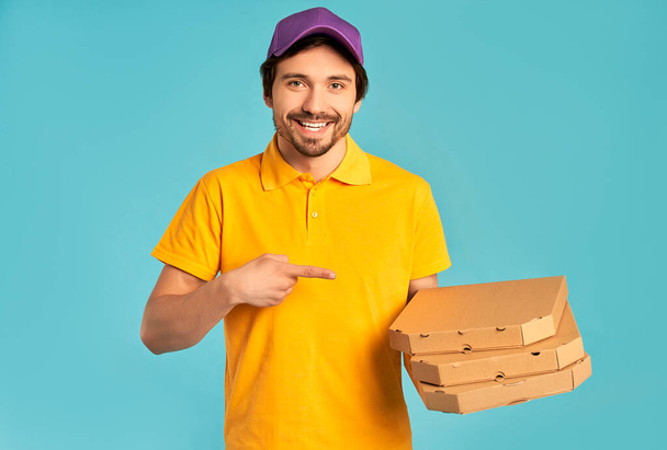 Młody kurier w mundurze pokazuje palec w pudełkach po pizzy odizolowanych na niebieskim tle. Dostawa do domu. - Zdjęcie, obraz