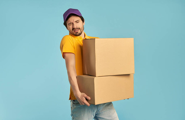 Young bearded courier delivery man in uniform holding heavy boxes isolated on blue background. Home delivery. - Photo, Image