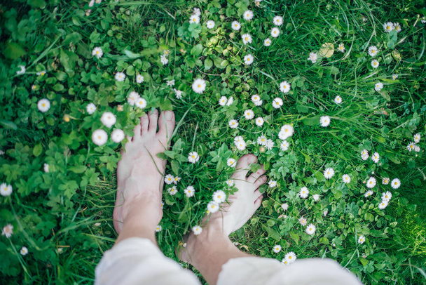 Αρσενικά γυμνά πόδια σε πράσινο γρασίδι. Κλείσιμο των ποδιών σε φρέσκα λαχανικά και λουλούδια του καλοκαιριού. - Φωτογραφία, εικόνα