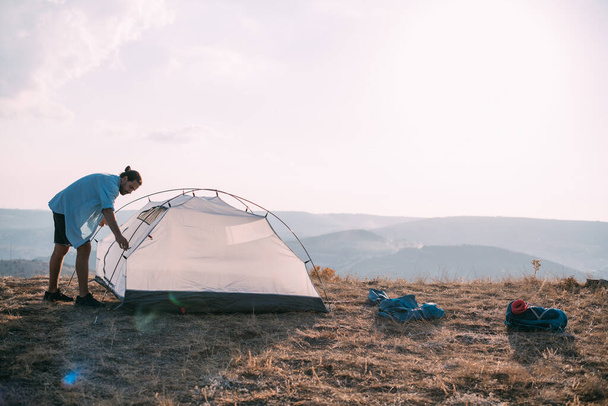 Mladý turista postaví stan v horách. Ten chlap si založil tábor na túře, sám sbírá stan v horách při západu slunce - Fotografie, Obrázek