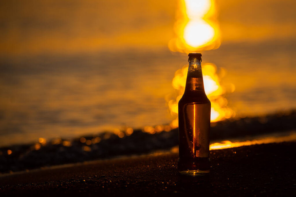 An ice cold beer / soda bottle standing straight on the golden sands at sunset. Summer evening - Photo, Image