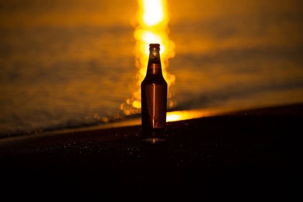Egy jéghideg sör / szódásüveg, ami egyenesen az arany homokon áll naplementekor. Nyári este - Fotó, kép