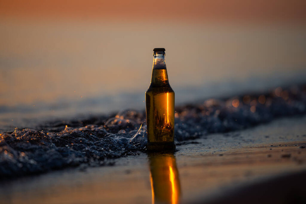 Gün batımında sahilde bir şişe light bira. Dalgalı deniz kıyısı. Bokeh arkaplanı. - Fotoğraf, Görsel