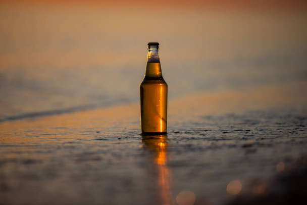 Una botella de cerveza ligera en la playa al atardecer. Orilla del mar con olas. Fondo de Bokeh. - Foto, Imagen
