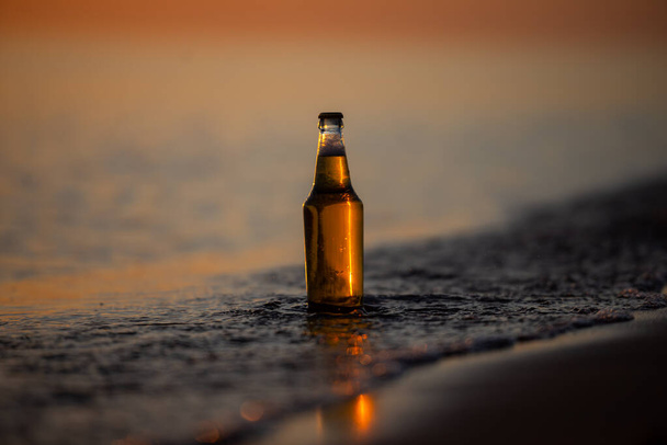 Egy üveg világos sör a parton naplementekor. Tengerpart hullámokkal. Bokeh háttér. - Fotó, kép