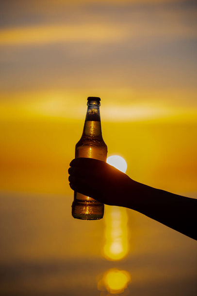 Mujeres sosteniendo una botella de cerveza con temática de verano en sus manos al atardecer. Botella de cerveza luz dorada del sol, formando burbujas bokeh. Profundidad superficial del campo - Foto, Imagen