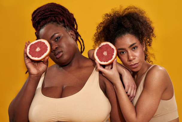 Two young african american women in underwear looking at camera, holding halfs of juicy grapefruit, posing together isolated over orange background - Fotó, kép