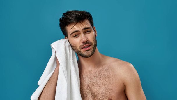 Studio portrait of confident handsome shaven man with stylish hairdo, using a white towel to wipe himself after shaving and taking a morning shower, posing isolated over blue background - Foto, Imagem