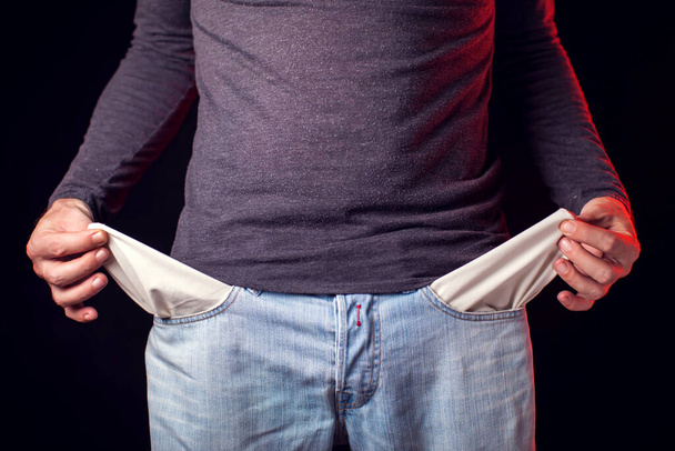 Мужчина показывает пустой карман своих джинсов на черном фоне. Концепция бедности и финансовых проблем - Фото, изображение