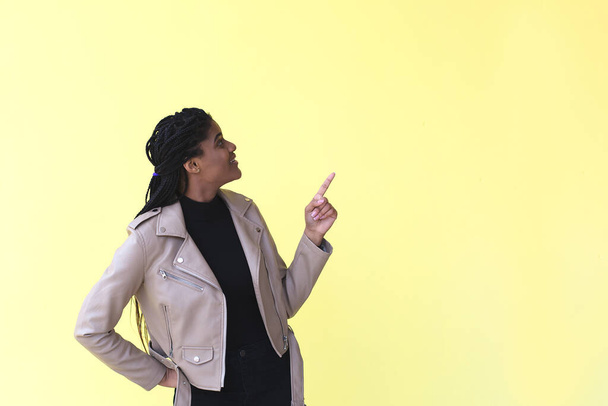 a beautiful and happy African-American woman points to a place for advertising, on a yellow background. - 写真・画像
