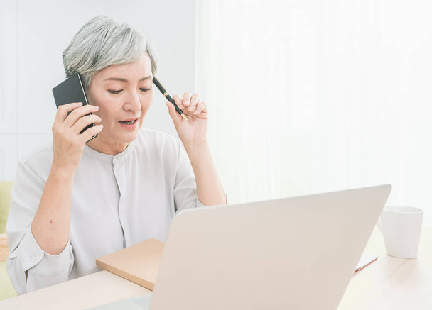 Asian senior woman at phone works on her laptop at home. - Photo, image