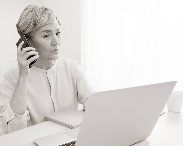 Asiatisch senior frau bei telefon works auf sie laptop bei zuhause. - Foto, Bild