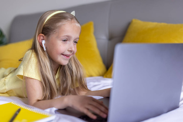 Cute little caucasian girl with blonde hair in fashionable dress illuminating yellow color sitting at home during coronavirus pandemic quarantine and using laptop. Stay at home during covid-19 - Photo, image