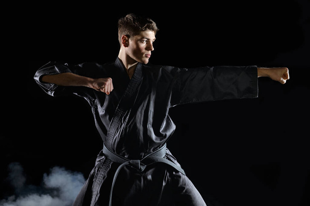 Male karate fighter in black kimono, combat stance, dark background. Man on workout, martial arts, fighting competition - Photo, Image