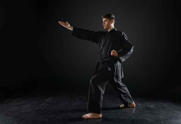 Male karateka, fighter practice in black kimono, combat stance, dark background. Man on workout, martial arts, training before fighting competition - Photo, Image