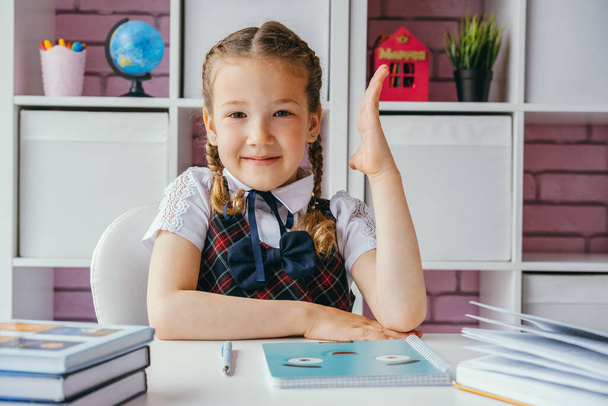 Beautiful little school girl in class is sitting at table with raised hand - Photo, image