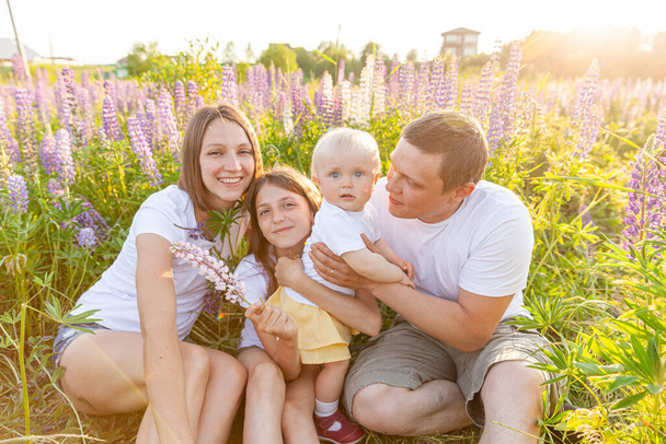 Happy family mother father embracing kids outdoor. Woman man baby child and teenage girl sitting on summer field with blooming flowers background. Happy family mom dad and daughters playing on meadow - Photo, Image