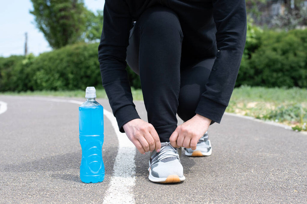 Одна голубая пластиковая бутылка изотонического напитка и спортсменка, завязывающая шнурки на кроссовках на беговой дорожке, крупным планом. - Фото, изображение