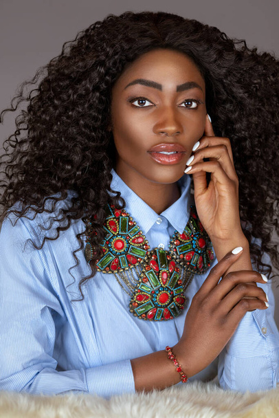 Porträt einer sinnlichen jungen schwarzen Frau mit langen lockigen schwarzen Haaren und schönem Make-up, die allein auf Fell in einem Studio mit grauem Hintergrund sitzt und ein blaues Hemd und bunten Schmuck trägt. - Foto, Bild