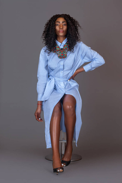 Portret van een sensuele jonge zwarte vrouw met lang krullend zwart haar en mooie make-up zitten op een barstoel door haarzelf in een studio met een grijze achtergrond dragen van een blauwe jurk en kleurrijke sieraden. - Foto, afbeelding