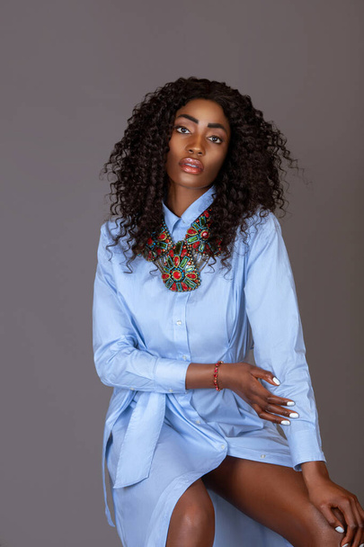 Porträt einer starken jungen schwarzen Frau mit langen lockigen schwarzen Haaren und schönem Make-up, die allein auf einem Barstuhl in einem Studio mit grauem Hintergrund sitzt und ein blaues Kleid und bunten Schmuck trägt. - Foto, Bild