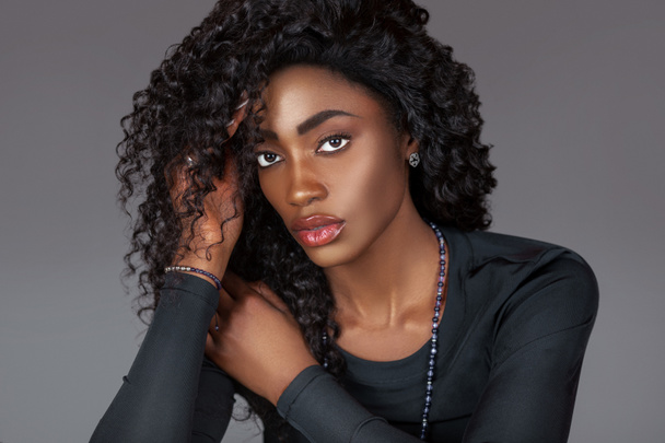 Retrato de uma bela jovem negra com cabelo preto encaracolado longo, bela maquiagem sentada sozinha na pele em um estúdio com um fundo cinza vestindo uma camisa de manga longa preta e jóias. - Foto, Imagem