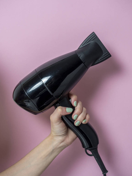 Pembe arka planda bir kadının elinde siyah saç kurutma makinesi. Saç bakım ürünleri. Dikey fotoğraf - Fotoğraf, Görsel