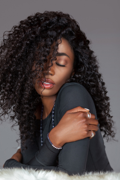 Portrait of a sensual young black woman with long curly black hair, beautiful makeup sitting by herself on fur in a studio with a grey background wearing a black long sleeve shirt and jewelry. - Foto, Imagem